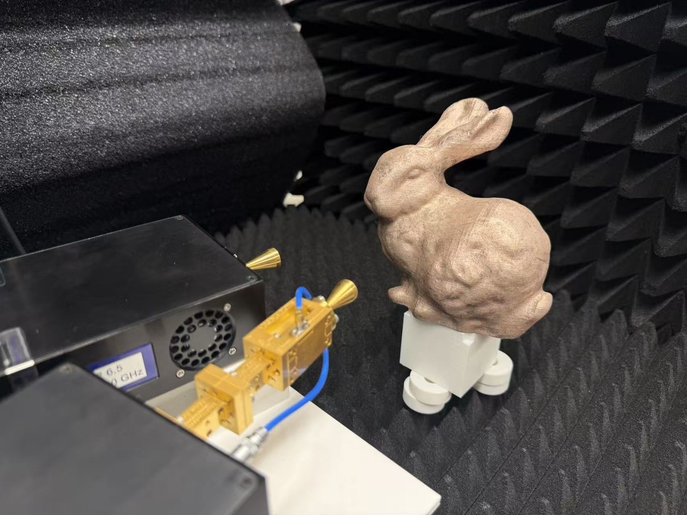 A 3D-printed bunny coated in conductive ink, on a narrow mount inside absorber foam next to two horn antennas.